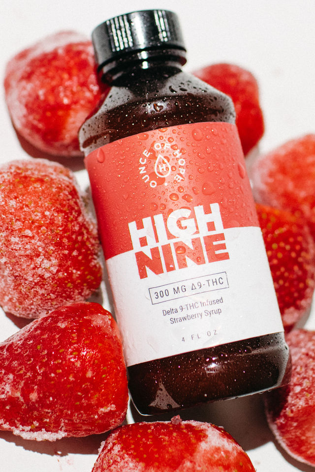 Close-up of a brown glass bottle of Delta-9 THC strawberry syrup with a pink label and water droplets, nestled among frozen strawberries on a white background.