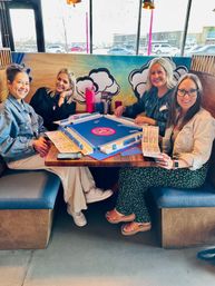 Four women smiling in a bright cafe booth playing a blue tabletop mahjong-style game, colorful cloud mural behind them and large windows showing parked cars and a shopping strip outside.