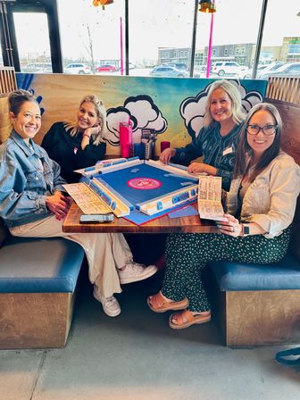 Four women smiling in a bright cafe booth playing a blue tabletop mahjong-style game, colorful cloud mural behind them and large windows showing parked cars and a shopping strip outside.