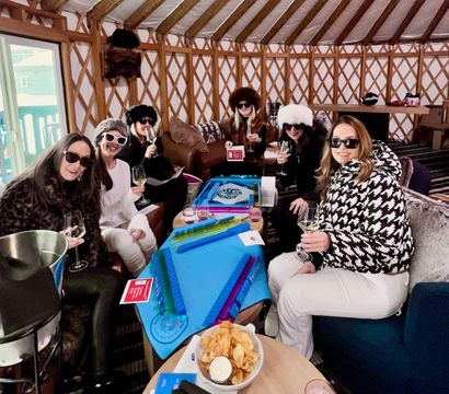Group of women in fur hats and sunglasses enjoying wine and champagne inside a cozy yurt-style ski lodge, gathered around blue tabletop games with chips and a snowy balcony view.