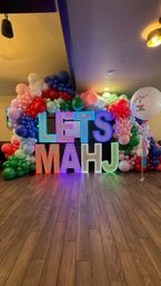Bright neon letters reading LETS MAHJ framed by a playful multicolor balloon arch and a large celebratory balloon on hardwood floor — festive indoor event decor