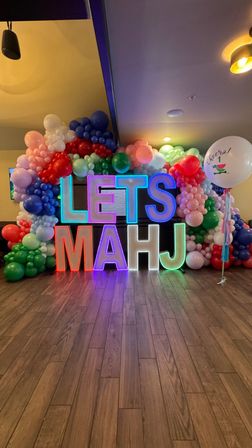 Bright neon letters reading LETS MAHJ framed by a playful multicolor balloon arch and a large celebratory balloon on hardwood floor — festive indoor event decor