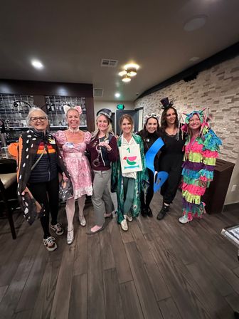 Group photo of seven women in colorful costumes at an indoor party venue — butterfly cape, pink frilly dress, police hat with handcuffs, peacock robe with number tag, blue musical-note cutout, black top-hat outfit, and multicolored fringe piñata suit.