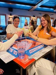 Smiling friends toasting frozen margaritas over a colorful tabletop mahjong game in a cozy bar with wooden beams and a blue ceiling.