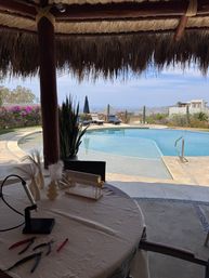 Thatched palapa patio with a round table of craft tools and displays, overlooking a curving pool, lounge chairs, cacti and a distant ocean view — sunny coastal getaway.