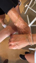 Close-up of three wrists wearing layered gold chain bracelets and a sparkling diamond tennis bracelet, casual sneakers and boutique floor in the background — jewelry styling shot