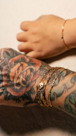 Close-up of a tattooed forearm with a colorful rose floral tattoo and layered gold chain bracelets next to a baby’s hand wearing a delicate gold bracelet