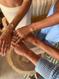 Sunlit hands stacked in a friendship pose, showcasing delicate gold chain and shell bracelets, rings and colorful manicures over a round wooden table — casual summer jewelry styling.