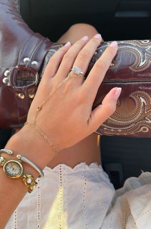 Close-up of a hand with French manicure and delicate gold hand-chain draped over an embossed brown leather cowboy boot; silver rectangular ring, gold watch bracelet and white eyelet skirt suggest western summer accessories.