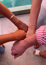 Four wrists joined in a fist-bump showing delicate gold chain friendship bracelets with tiny pearls, poolside tiles and sandals in the background