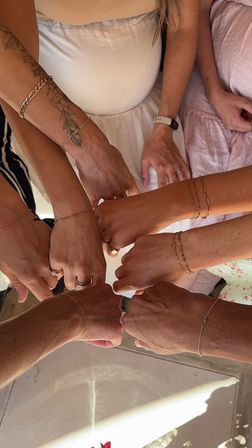 Overhead close-up of a circle of hands and fists joining in a friendly fist-bump, featuring delicate chain bracelets, rings, a floral tattoo, and pastel summer outfits.