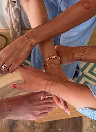 Several wrists and a foot wearing delicate gold chain bracelets, a shell bangle, rings and pink manicures arranged casually over wood and patterned tile — stylish summer jewelry close-up.