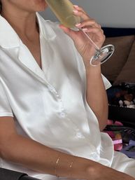 Close-up lifestyle shot of a person in white satin pajamas sipping champagne from a flute, showing manicured nails and delicate gold bracelets — relaxed at-home morning vibe.
