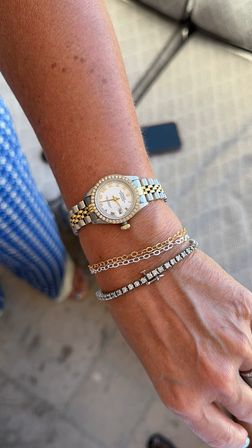 Close-up of a sunlit wrist wearing a two-tone gold-and-silver watch with a diamond bezel and two bracelets — a delicate gold chain and a sparkling tennis bracelet, patio tiles and blue patterned fabric in background.
