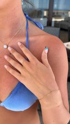 Close-up of sun-kissed person's hand resting on chest wearing a blue bikini top, delicate gold hand-chain bracelet and pendant necklace, with French-tip nails and turquoise accent in a sunny poolside patio setting.