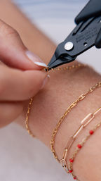 Close-up of hands using black pliers to fasten a delicate gold chain bracelet on a wrist, showing layered gold chains and tiny red bead accents — artisan jewelry repair.