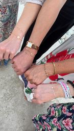 Four women's wrists displaying stacked bracelets, beaded bracelets and a gold watch over colorful floral dresses on a light stone patio — stylish summer accessories.