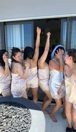 Bridal party in matching silk pajamas with the bride in a white slip and veil, laughing and dancing on a resort-style balcony by sliding glass doors and a lava-rock fire pit.