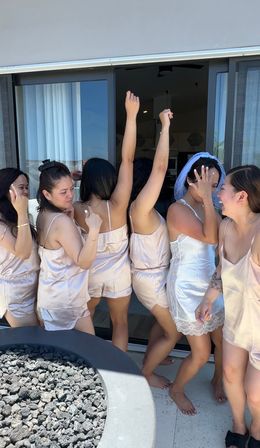 Bridal party in matching silk pajamas with the bride in a white slip and veil, laughing and dancing on a resort-style balcony by sliding glass doors and a lava-rock fire pit.
