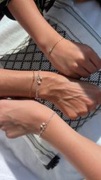 Three sun-kissed wrists wearing delicate gold and silver chain bracelets with tiny pearl charms on a black-and-white patterned towel.