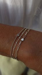 Close-up of minimalist silver bracelet stack on tanned wrist — delicate beaded and paperclip chains with a tiny heart charm and small crystal accent.