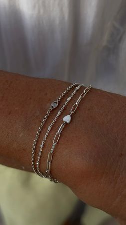 Close-up of minimalist silver bracelet stack on tanned wrist — delicate beaded and paperclip chains with a tiny heart charm and small crystal accent.
