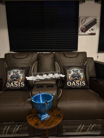 Cozy indoor cigar-lounge scene: brown leather reclining seats with matching illustrated dog-and-palm pillows, a tray of whiskey glasses, blue ash bowl on a wooden side table, black blinds and cigar wall art.