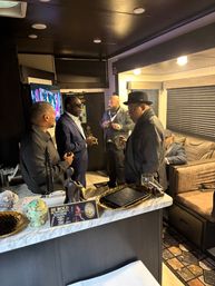 Group of well-dressed men in suits and hats enjoying cigars and conversation in an upscale VIP lounge inside a private RV, marble countertop with decorative tray and a 'Be Bold' sign in the foreground.