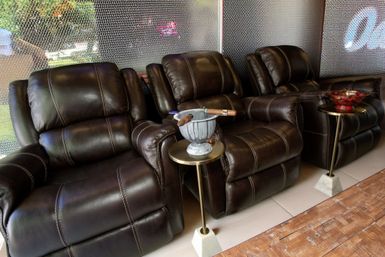 Three plush dark brown leather recliners in a lounge area by a screened window, with small side tables holding ashtrays and cigars