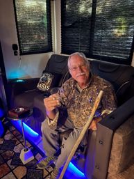 Smiling senior man with glasses and a mustache seated in a leather recliner in a cozy dimly lit lounge, holding a plastic cup and a wooden walking stick, with blue LED accent lighting and a tropical-themed throw pillow nearby.