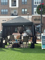 Cozy black canopy gazebo with sheer curtains, four chairs and a small table on a rug over artificial turf in a downtown plaza with brick buildings and a holiday wreath