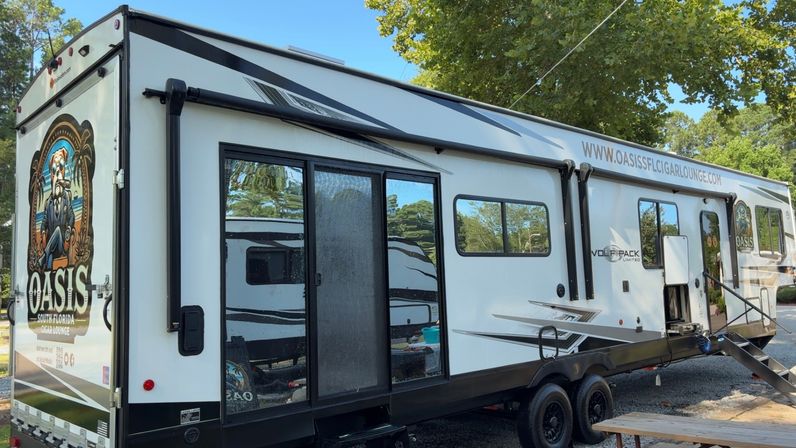 White travel trailer RV with black trim and retractable awning parked under trees in a sunny Florida campground; sliding glass door, fold-out metal steps and twin axles, with a colorful tropical cigar-lounge themed mural on the rear panel.