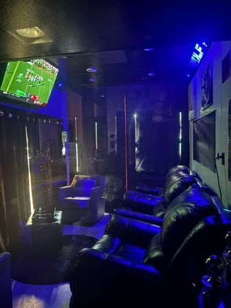 Home theater lounge with black leather recliners in a row, blue LED accent lighting, wall-mounted TV showing a football game, cozy armchair with ottoman, dark curtains and modern entertainment setup.