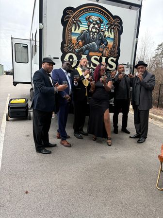 Seven adults in formal and cocktail attire toasting with drinks while posing in front of a decorated event trailer featuring a stylized dog emblem, parked in an outdoor lot.