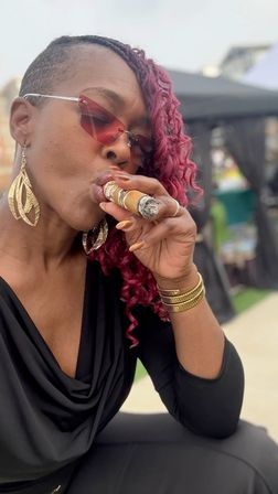 Stylish person with a half-shaved head and curly magenta braids wearing red-tinted sunglasses and gold leaf earrings, dressed in black and smoking a cigar at an outdoor market-style event.