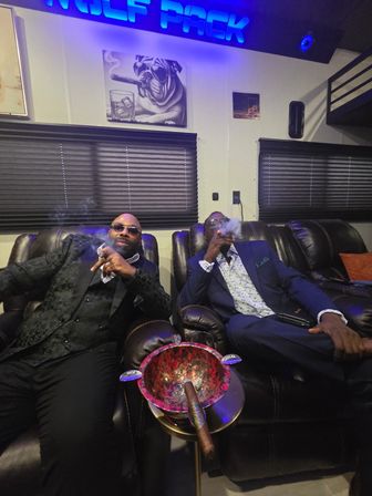 Two men in suits reclining on leather chairs in a dim cigar lounge, exhaling smoke under a blue neon sign with a large ashtray and cigars in the foreground and a bulldog cigar art print on the wall.