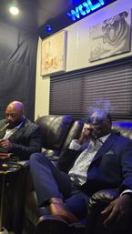 Two well-dressed men in suits relaxing on leather recliners in a dim cigar lounge, smoking cigars with smoke curling up, blue neon sign and playful wall art above, small side table with a glass nearby