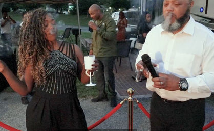 Outdoor evening party scene: woman in a beaded black dress holds a white goblet and gestures while a bearded man in a white shirt holds a microphone and cigar near red velvet rope stanchions, guests mingling in the background.