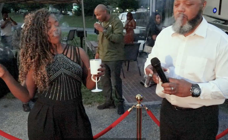 Outdoor evening party scene: woman in a beaded black dress holds a white goblet and gestures while a bearded man in a white shirt holds a microphone and cigar near red velvet rope stanchions, guests mingling in the background.
