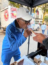 Person in blue tracksuit and white cap taking a puff from a cigar handed to them at an outdoor city street market stall, vendor table with boxes and mug and a delivery truck and decorated storefronts in the background.