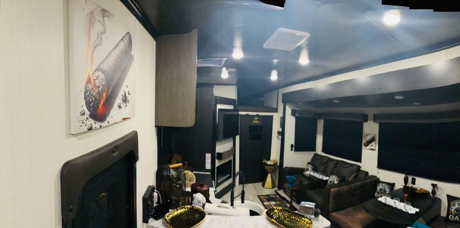 Modern travel trailer interior with a compact kitchen island and sink, gold decorative bowls, cozy leather sofa, dark window blinds, recessed ceiling lights, and framed wall art.