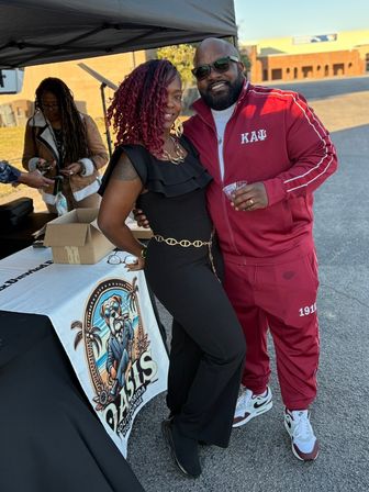 Smiling couple posing at an outdoor vendor tent during a parking-lot community event — woman with red braids in a black jumpsuit and gold chain belt, man in a maroon tracksuit and sunglasses holding a drink, colorful vendor table and people in the background.