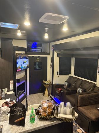 Cozy RV lounge interior with leather theater recliners, wall-mounted TV showing a basketball game, a VIP ONLY door, snack trays and cups on a marble-look counter, cigar bar sign, and blue LED accent lighting.