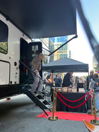 Man stepping down stairs of a white RV with a black awning at a downtown street festival, red carpet and gold stanchions leading to a vendor pop-up tent with boxes on a black table, glass skyscrapers and city crowd in the background.