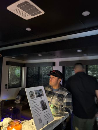 Dimly lit modern lounge reception with marble-pattern countertop, a man in a plaid shirt and cap reading a menu on a display stand, glass tip jar and candle on the counter, blurred person and closed blinds in the background.