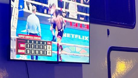 Wall-mounted TV broadcasting a punch-filled boxing match in a bright blue ring, two fighters and a referee visible, on-screen unofficial scorecard showing totals 46 and 49.