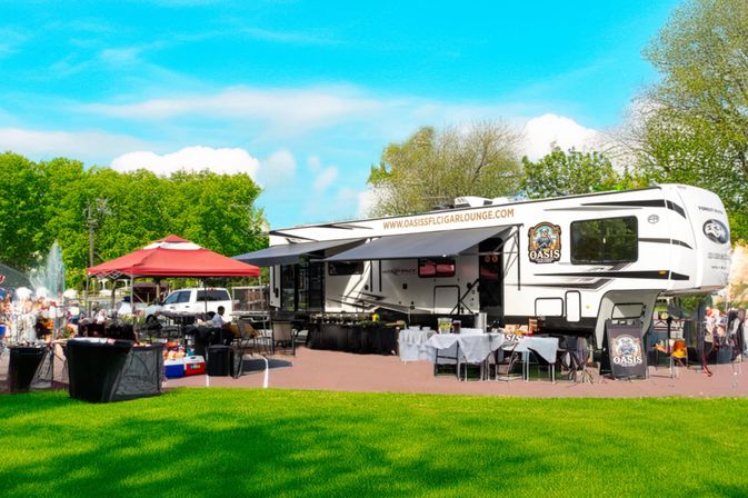 White fifth-wheel RV converted into a pop-up outdoor lounge with awning, tables and a red canopy at a sunny park festival beside a fountain and green lawn.
