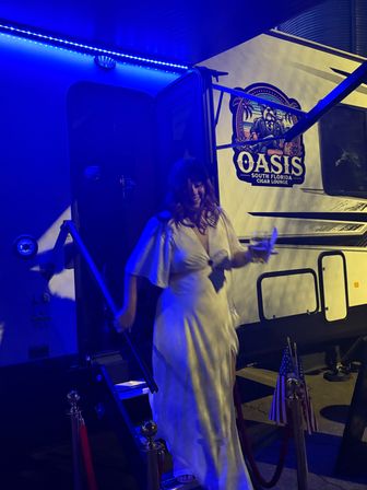Woman in a white dress stepping down from an illuminated trailer at a nighttime outdoor lounge event in Florida, bathed in blue LED light, holding a drink with velvet ropes and small American flags nearby.