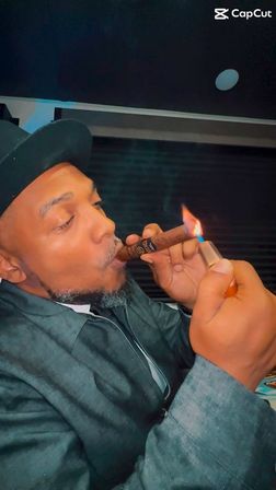 Close-up of a person in a dark hat and jacket lighting a cigar with a handheld lighter, warm flame glowing against a dim indoor background, relaxed cozy moment.