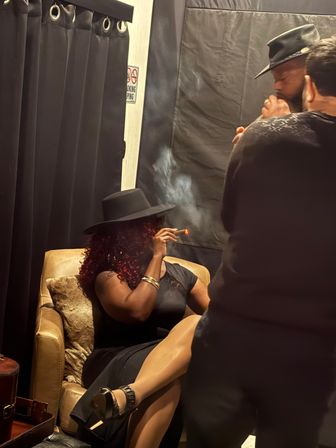Person in black dress and wide‑brim hat reclining in a leather armchair, smoking a cigar in a dim, smoky indoor cigar lounge with two standing companions.